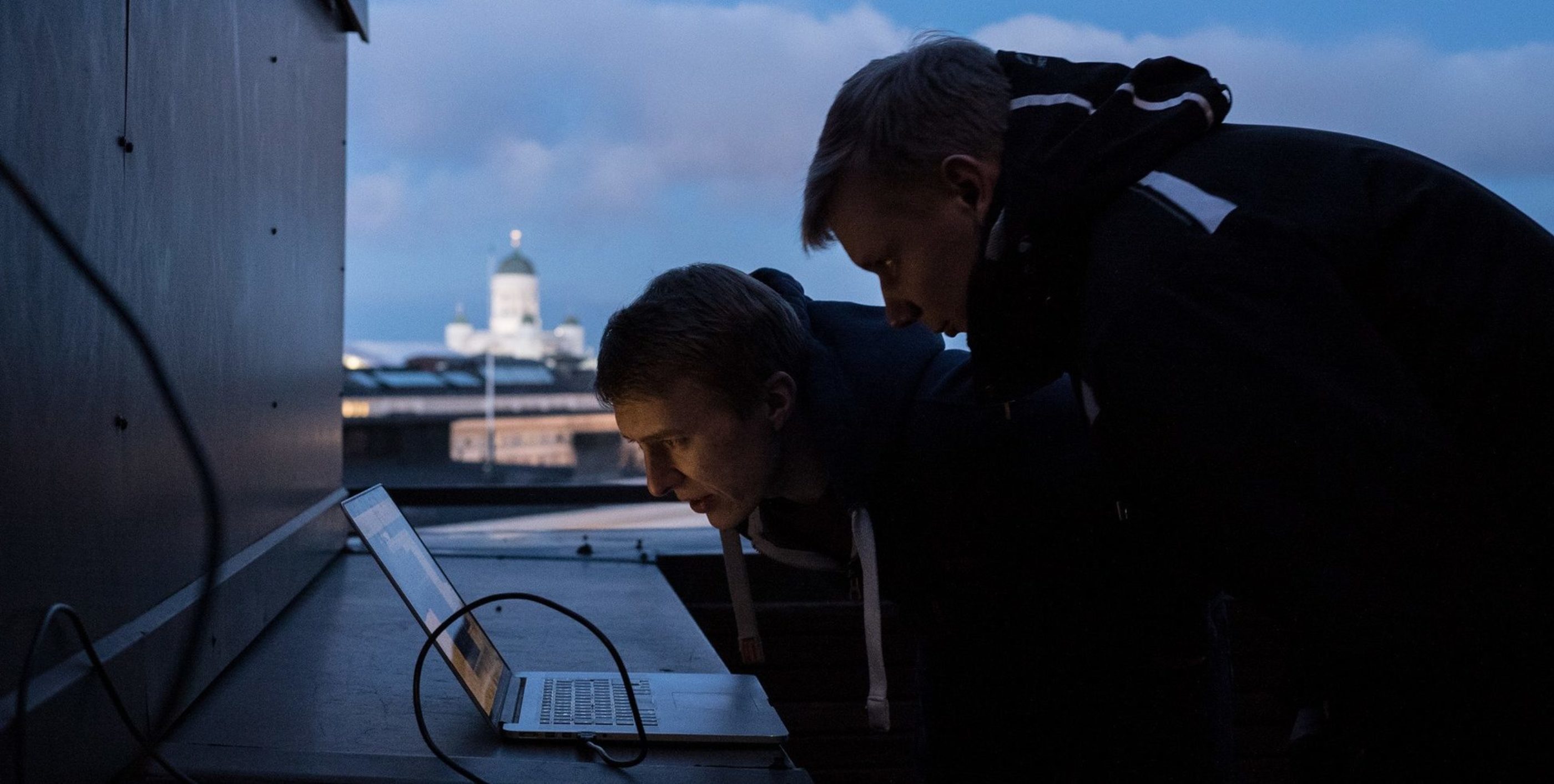 Two men staring at a computer screen from a close distance at an evening. Behind them, the skyline of Helsinki and the evening sky with some clouds.